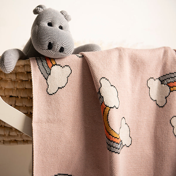 Knitted hippo toy doll on a light pink cotton blanket with cloud and rainbow patterns.