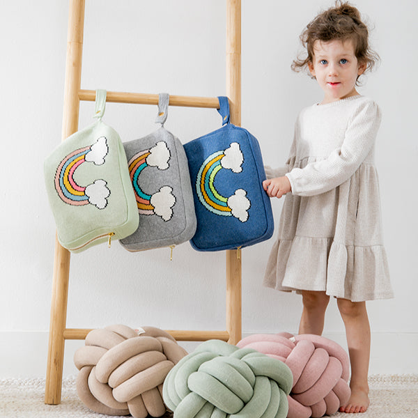 Child holding a blue blanket pouch with rainbow design next to multiple colourful knot pillows on a wooden ladder.