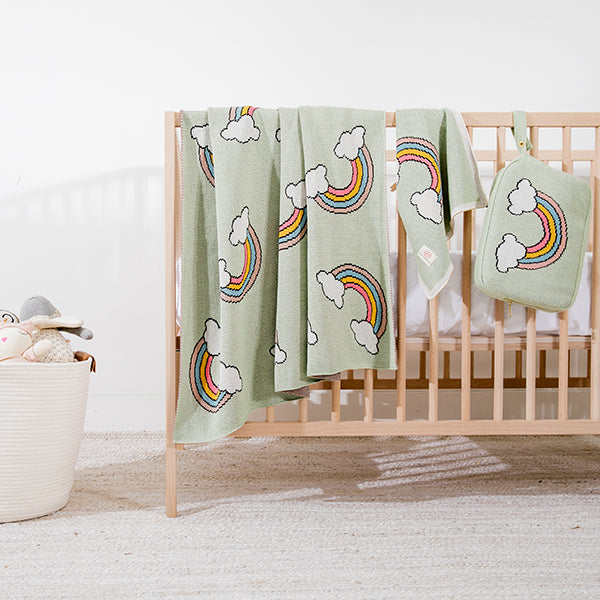 Mint green baby blanket, blanket pouch and burp cloth with rainbow and cloud patterns draped over a crib in a nursery.