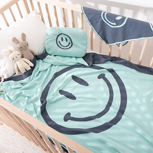 Children's crib with mint blue smiley face cotton baby blanket and pouch, two bunny plush toys, and a cloth.
