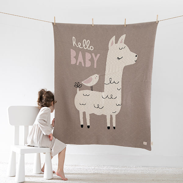 Taupe organic cotton blanket with llama design and 'Hello Baby' text, hanging on a line with a child sitting next to it on a white chair.