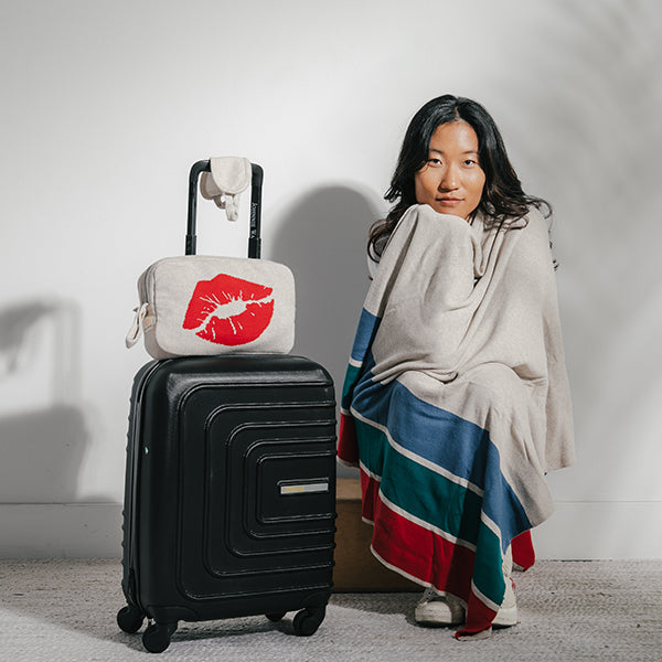Woman wrapped in a beige striped blanket next to a suitcase with a small bag featuring red lips on a plain background