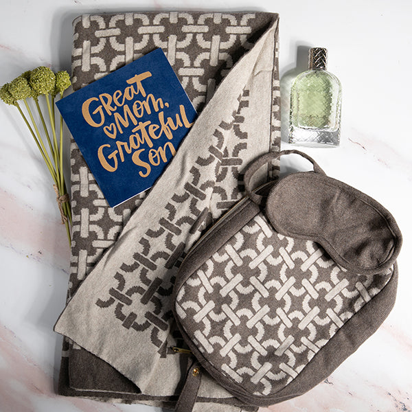 Set of chain link patterned blanket, brown eye mask, and travel cologne on a white marble surface