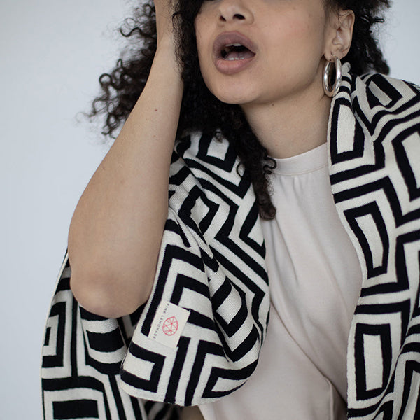 Woman wearing a black and white geometric patterned throw with a plain background.