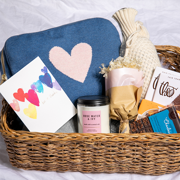 A variety of items including a blue and pink reversible blanket set, a zip pouch, a wristlet loop, a sleep mask, a candle, a card with a heart design, displayed in a wicker basket.