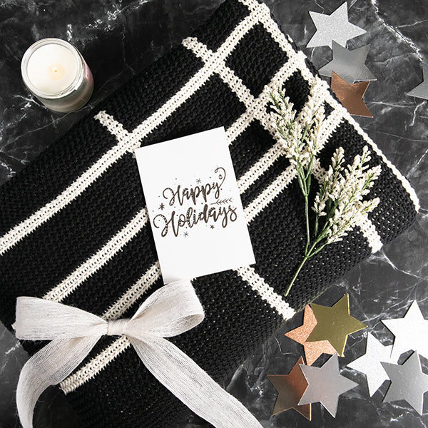 Gift wrapped black and white moss-knit throw blanket with a 'Happy Holidays' card on a dark surface with decorative stars.