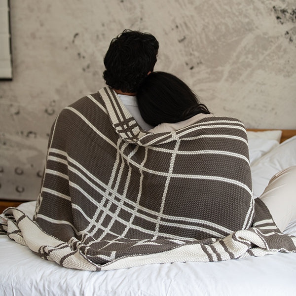 Couple wrapped in a double moss knit dark brown cotton blanket on a bed with a textured wall in the background.