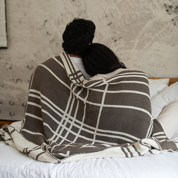 Couple wrapped in a double moss knit dark brown cotton blanket on a bed with a textured wall in the background.