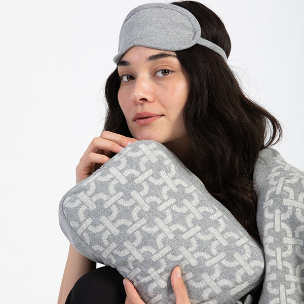 Woman wearing a gray sleep mask and holding a matching gray pouch with a chain link pattern on a white background.