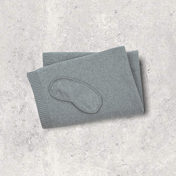 Gray cotton travel blanket with a cotton sleep mask on a textured gray surface.