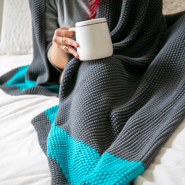 Person wrapped in a gray and teal moss-knit textured throw blanket holding a white mug.
