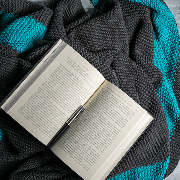 Open book placed on a moss knit blue and dark gray cotton throw blanket.