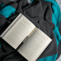 Open book placed on a moss knit blue and dark gray cotton throw blanket.