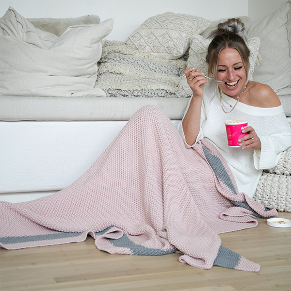 Woman sitting next to a white couch with a pink moss stitch knit cotton blanket, eating ice-cream from a pink cup.