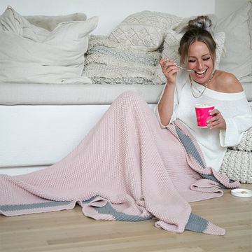 Woman sitting next to a white couch with a pink moss stitch knit cotton blanket, eating ice-cream from a pink cup.