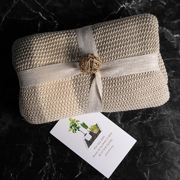 Beige woven moss knit cotton throw folded with a decorative ribbon on a dark textured surface next to a white gift card.