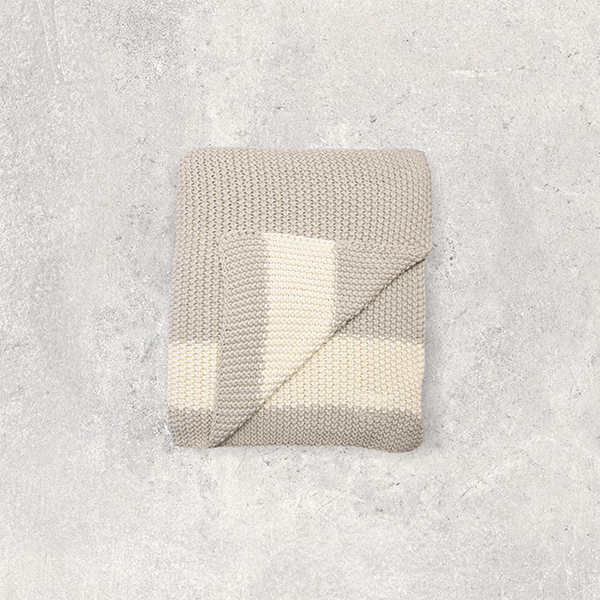 Folded beige and white moss knit cotton throw blanket on a textured gray surface.