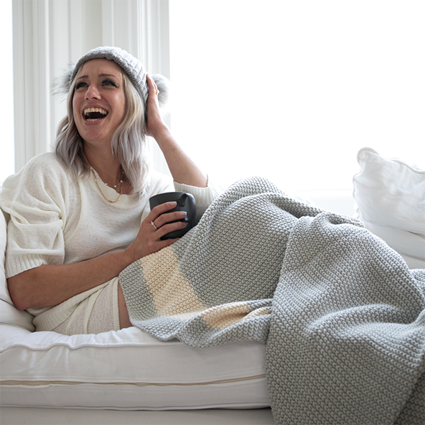 Woman sitting on a couch wrapped in a striped textured cotton throw, holding a mug and laughing.