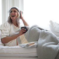Woman sitting on a couch wrapped in a striped textured cotton throw, holding a mug and laughing.