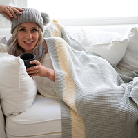 Woman sitting on a couch wearing a gray knit hat and wrapped under a moss-stitch vanilla and grey blanket, holding a mug.