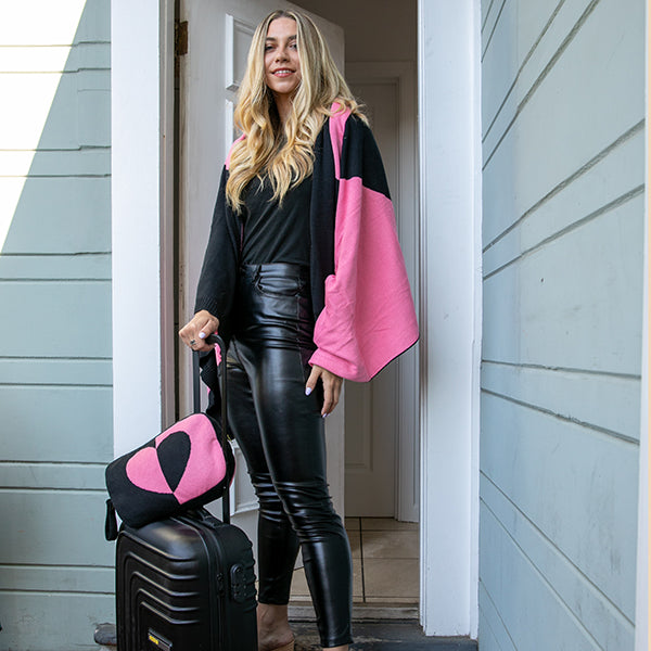 Woman holding a pink and black pouch on top of a suitcase, standing outside a house wrapped in a pink and black cotton blanket.