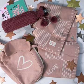 Pink travel blanket with heart design, matching pink pouch, and eye mask on a star-patterned background with a knitted wine bag.