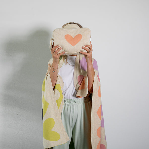 Person holding a beige bag with a heart design and wearing a blanket against a plain background.