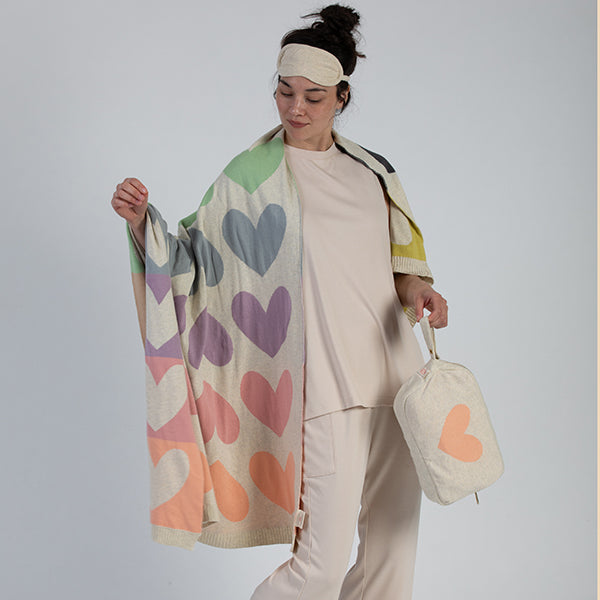 Woman wearing an eye mask and holding a colorful heart-patterned blanket and blanket bag against a plain background.