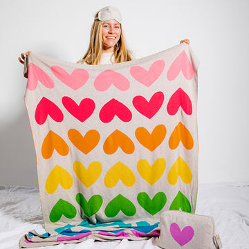Person wearing an eye mask and holding a colorful heart-patterned cotton blanket with a plain background.