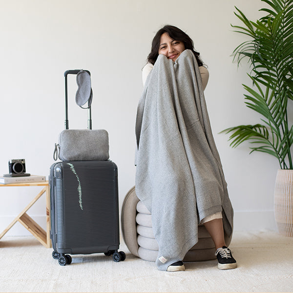 Woman wrapped in a gray textured reversible blanket next to a suitcase in a room with a plant.