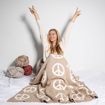 Woman sitting on a bed with a beige cotton throw blanket featuring peace signs, surrounded by decorative knot pillows.