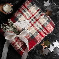 Red checked cotton throw with a candle, card, and decorative bow on a dark surface with star-shaped confetti.