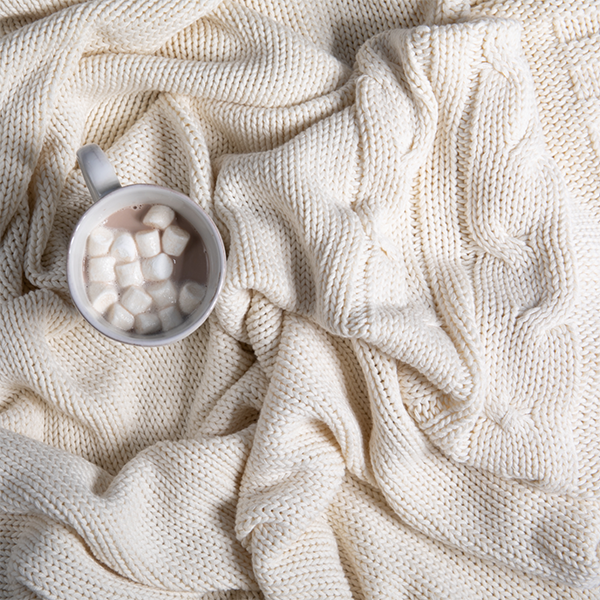 White mug of hot cocoa with marshmallows on a soft beige knitted cotton throw blanket.