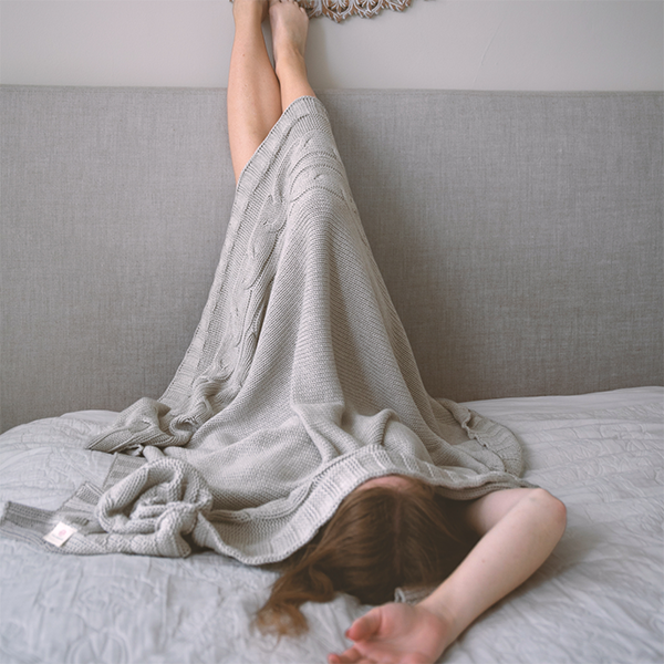Person lying on a bed with their feet on the wall while hiding under a soft beige textured cotton throw.