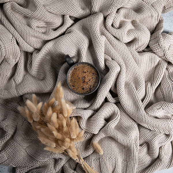 Cup of coffee and dried pampas grass placed on a textured beige cotton luxury throw blanket.