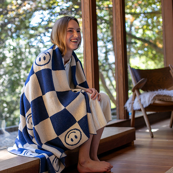 Woman sitting on an outdoor wooden bench with a blue and white checkered blanket featuring smiley face symbols.