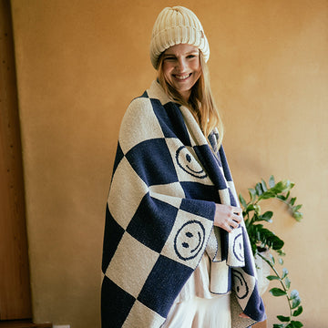 Woman wrapped in a navy blue and white checkered throw blanket with smiley face patterns, wearing a beige knit hat.