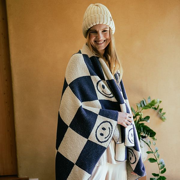 Woman wrapped in a navy blue and white checkered throw blanket with smiley face patterns, wearing a beige knit hat.