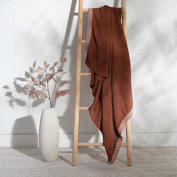Rust coloured textured blanket draped over a wooden ladder against a white wall with a vase and plant on the floor.