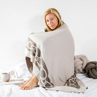 Woman sitting on a bed and wrapped in a large beige throw blanket with a dark brown and white chain pattern.
