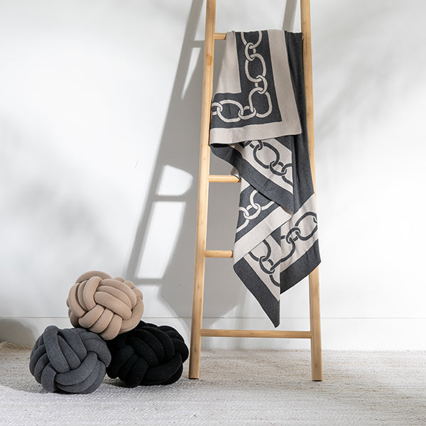 Dark grey cotton throw blanket with a chain design draped on a ladder next to knot cushions.