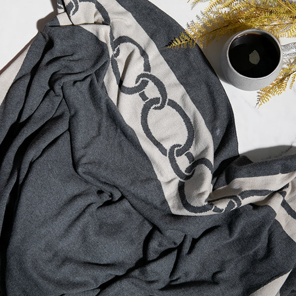 Dark gray throw with chain pattern draped over a surface, next to a cup of coffee and decorative elements.