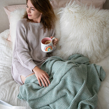 Woman sitting on a couch with a summer blue knitted throw blanket and a mug, holding a white mug with a rainbow design.