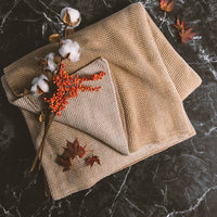Beige textured knitted throw blanket with decorative elements and flowers on a dark marble surface.