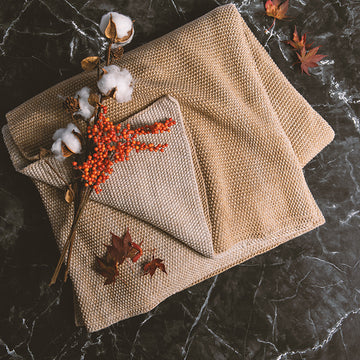 Beige textured knitted throw blanket with decorative elements and flowers on a dark marble surface.
