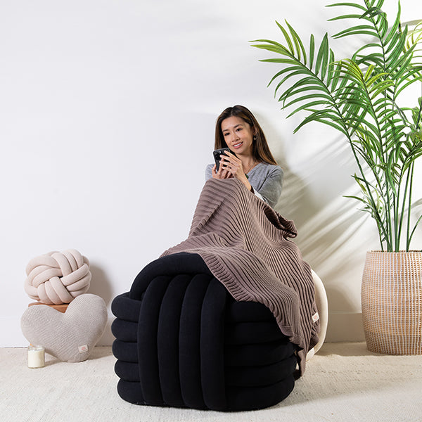 Woman sitting on a large knot cushion with a textured espresso brown throw blanket, holding a phone.