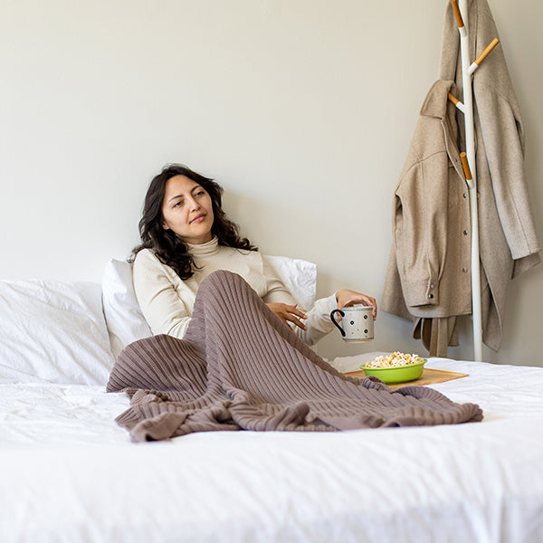 Woman sitting on a bed with a brown blanket, holding a mug and eating popcorn.