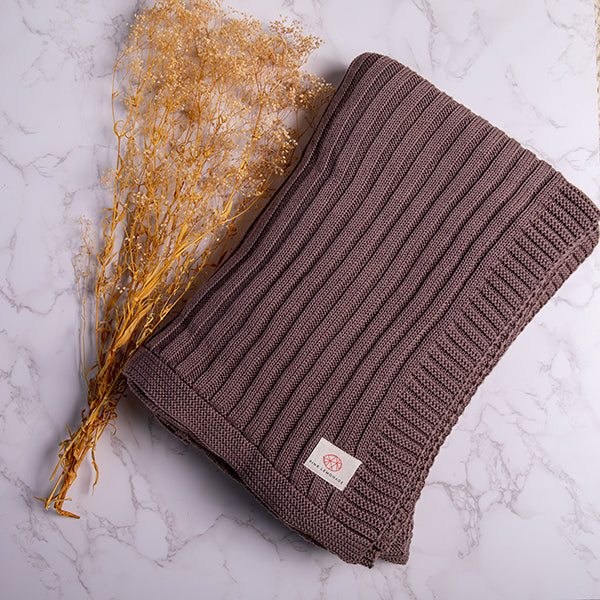 Espresso brown knitted blanket with a ribbed texture next to a dried plant on a white background.