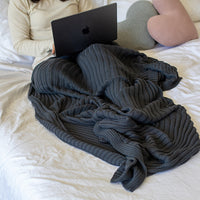 Person using a laptop under a charcoal gray and textured throw blanket on a bed.