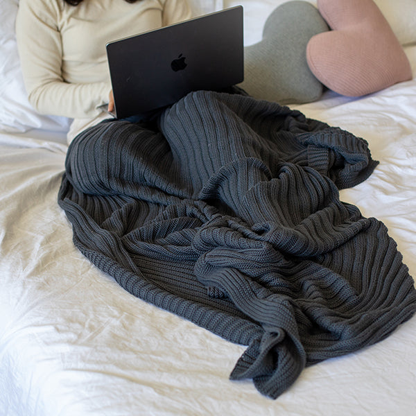 Person using a laptop under a charcoal gray and textured throw blanket on a bed.